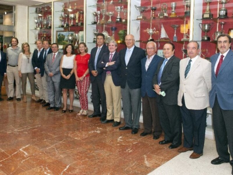 Grupo de personas posando en un salón con vitrinas de trofeos al fondo.