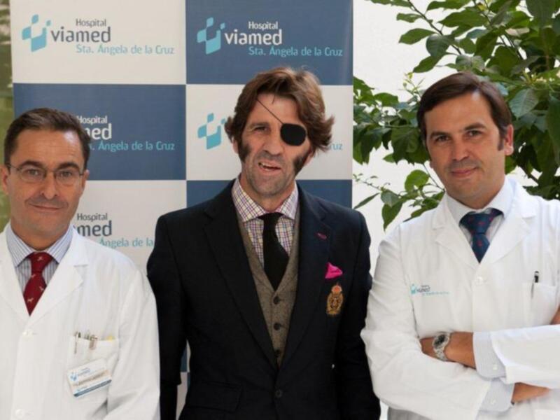 Dos profesionales de la salud posan frente a un fondo con el logotipo de Viamed Hospital Sta. Ángela de la Cruz, uno de ellos con una camisa y corbata, el otro con una bata de médico. La imagen sugiere un ambiente de trabajo en un hospital.