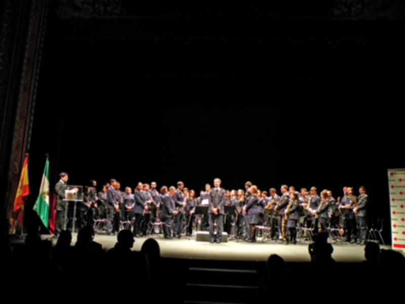 Concierto de banda en escenario iluminado, con músicos y bandera en el fondo.