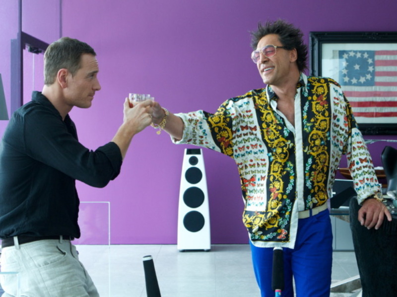 Un hombre en una chaqueta con estampado floral y pantalones azules celebra una copa con otro hombre en una sala con paredes de color púrpura y una bandera de los Estados Unidos en el fondo.