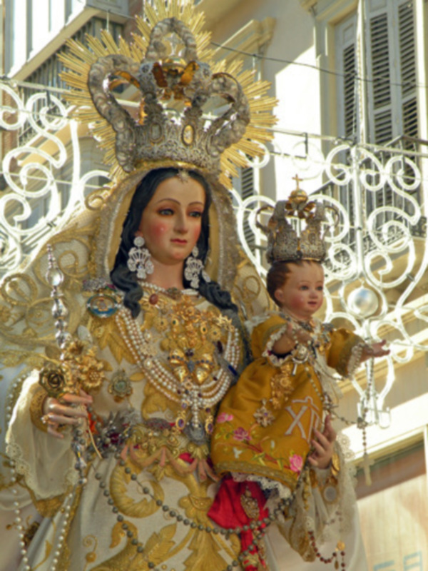Imponente imagen de la Virgen del Carmen, vestida con ornamentos dorados y una imagen del Niño Jesús en sus brazos, en un entorno festivo.