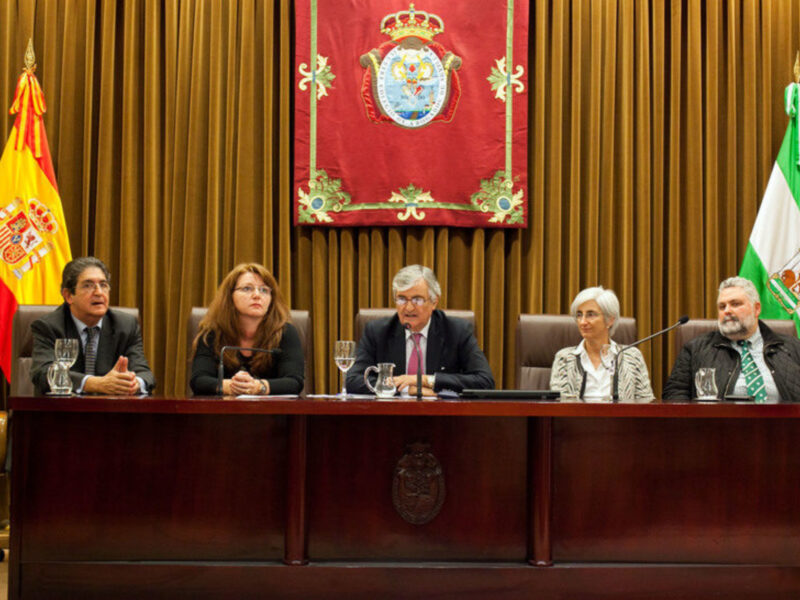 Una reunión formal con cuatro personas sentadas en una mesa frente a banderas y un escudo heráldico.