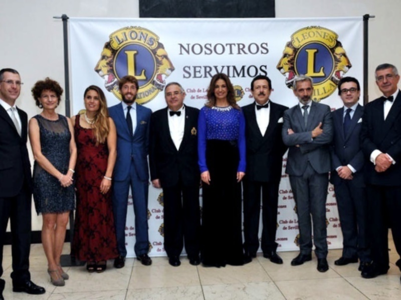 Conferencia del Club de Leones Internacional en Sevilla, con miembros vestidos de gala frente a un cartel con el lema 'Nosotros Servimos'.