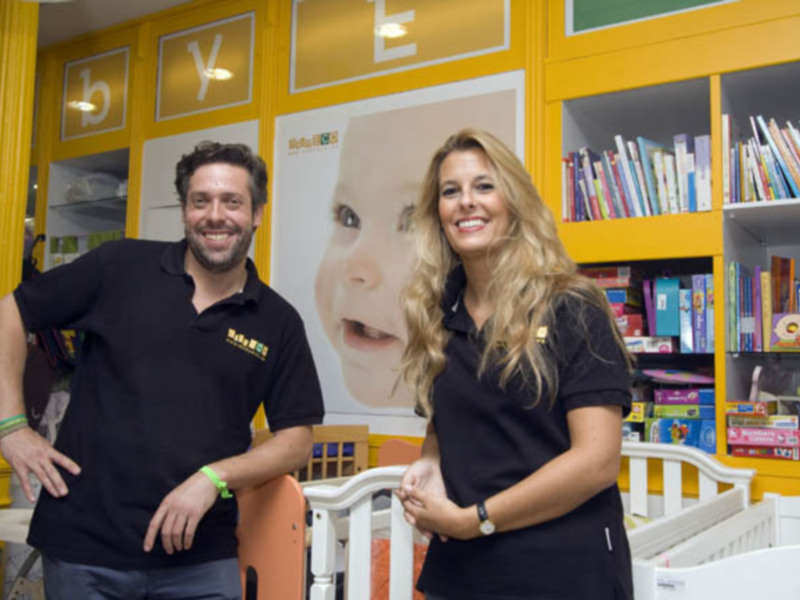 Dos personas sonrientes en una tienda infantil con estanterías llenas de libros y juguetes.