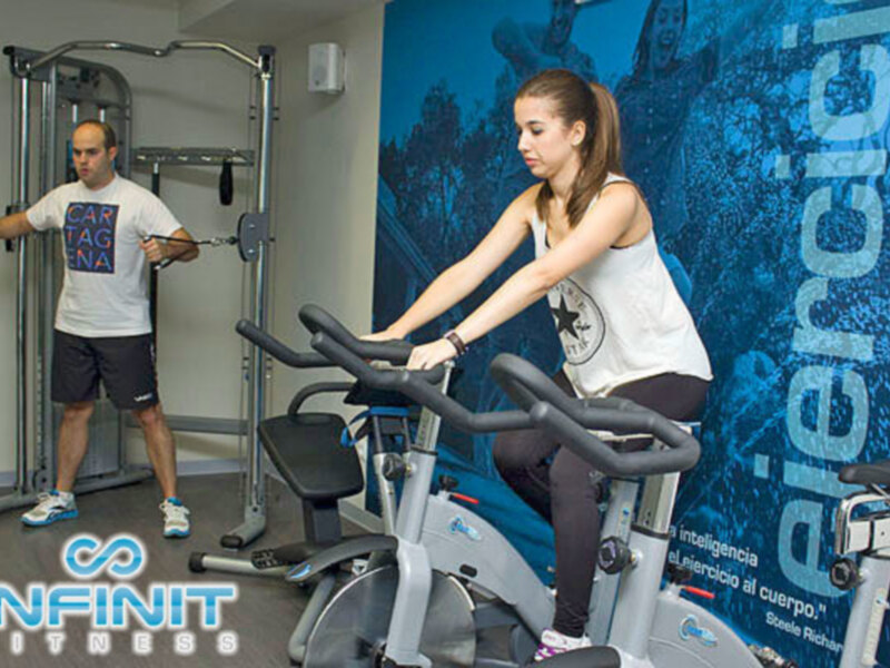 Persona realizando ejercicio en bicicleta estática mientras otro observa en un gimnasio con equipamiento de fitness.