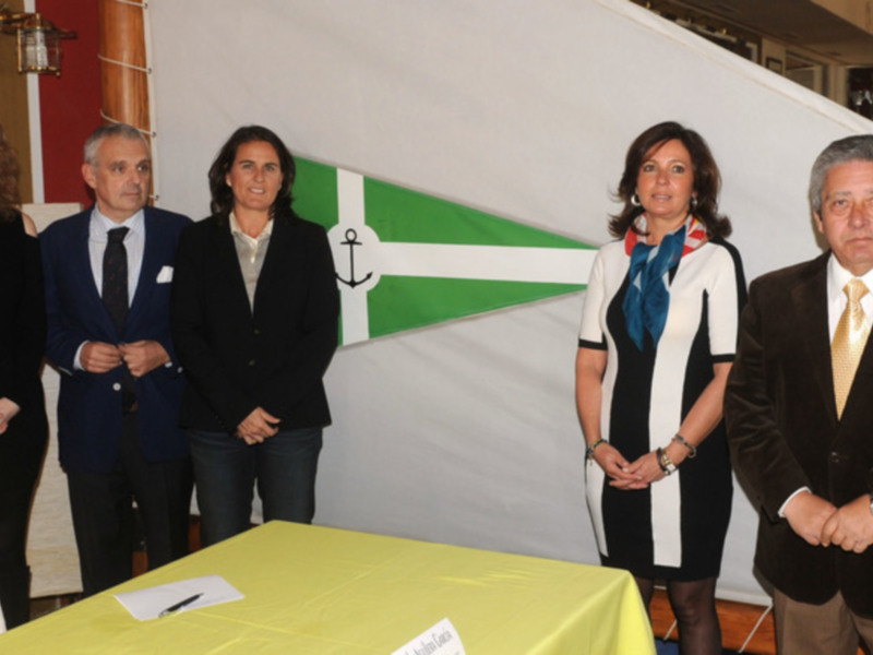 Grupo de personas en una reunión formal con una bandera de fondo y un escritorio con papel y bolígrafo delante.