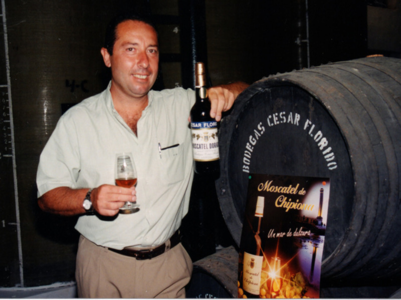 Hombre sosteniendo una botella de vino y un vaso, con barril y publicidad de Moscatel de Chipiona en el fondo.