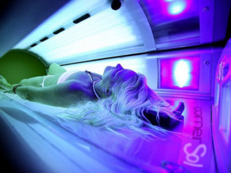 Una mujer con cabello largo y rubio está tumbada en una cabina de tanning con luces azules y púrpuras. La cabina tiene una etiqueta que dice 'Corner Spa'.