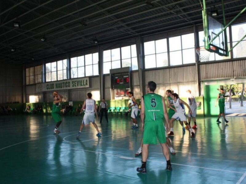 Partido de baloncesto en el Club Náutico Sevilla, con jugadores en acción en el aro y el marcador en el fondo.