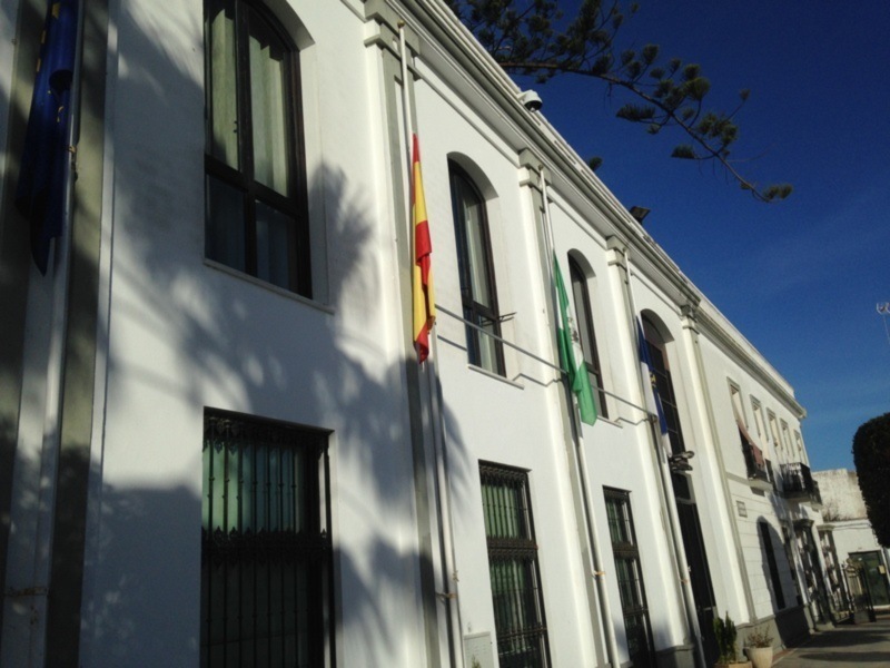 Fachada de edificio blanco con banderas española y andaluza, bajo un cielo azul claro.