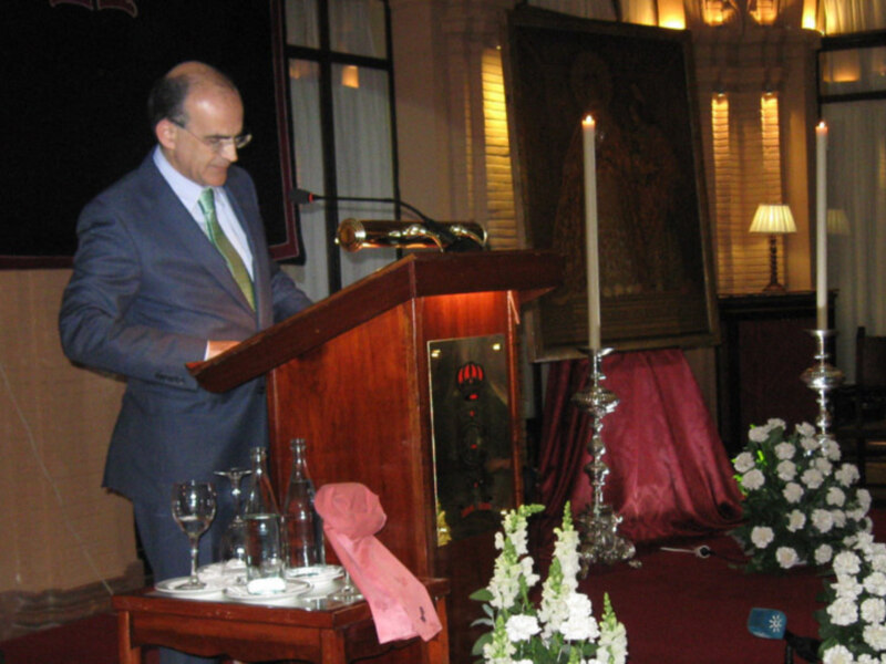 Hombre en traje negro leyendo en un podium con velas y flores blancas en el fondo.