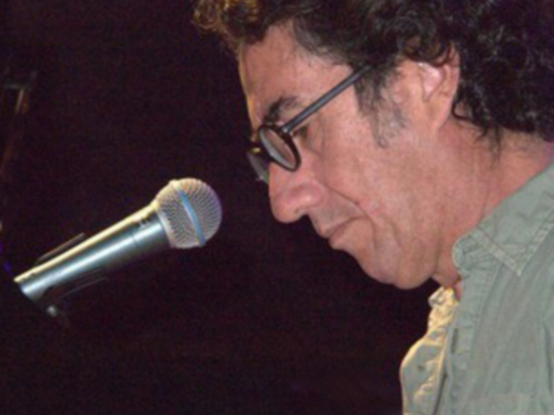 Un hombre con gafas y una camisa verde, posiblemente en una escena de concierto o actuación, con un micrófono en el frente.