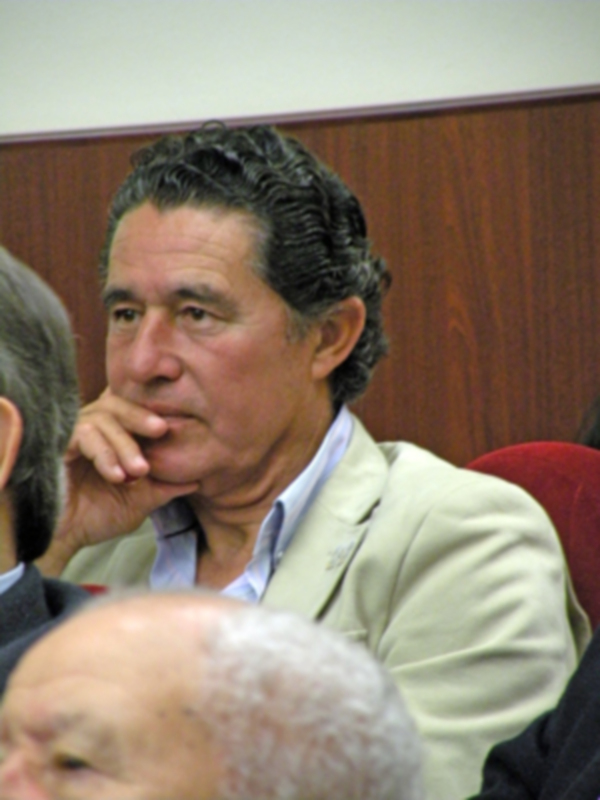 Hombre en una reunión o conferencia, con expresión pensativa y mano en la barbilla.