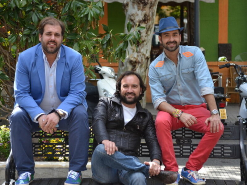 Tres hombres sentados en un banco en un entorno urbano, rodeados de vegetación y motocicletas.