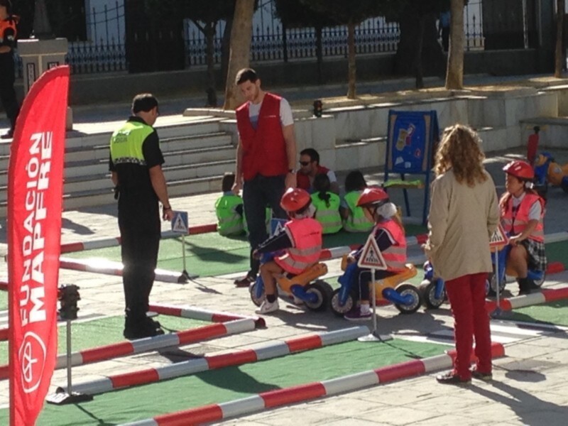 Niños aprendiendo sobre seguridad vial en un parque público, con adultos supervisando y banderas de Fundación Mapfre en el fondo.