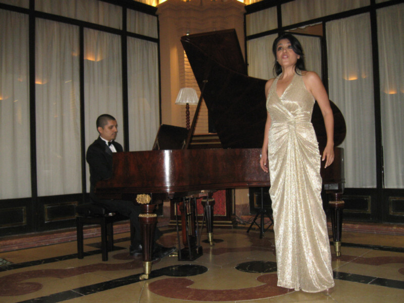 Una cantante en vestido de noche dorado se presenta junto a un pianista en un elegante salón de conciertos.
