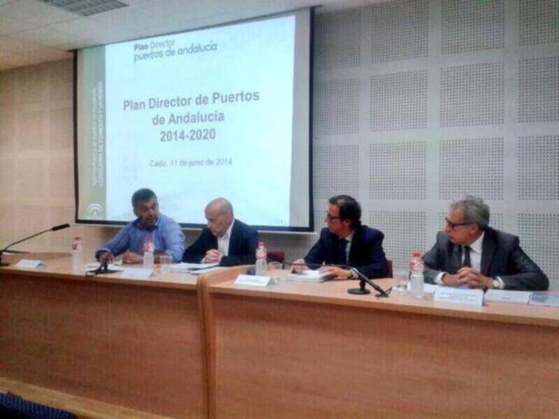 Presentación del Plan Director de Puertos de Andalucía en Cádiz, con cuatro personas en el escenario, frente a un proyector con la presentación en pantalla.