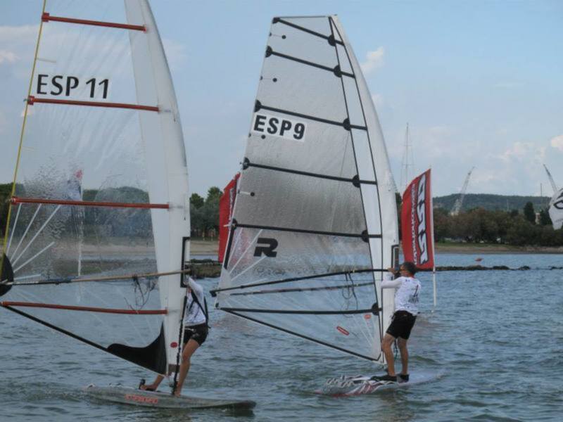 Dos personas practicando windsurf en el agua, con los palos y el casco visibles. La vela de uno de los windsurfers tiene la marca 'R'.