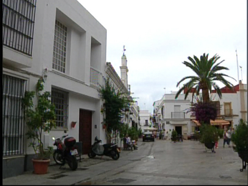 Calle con casas blancas y palmeras, motocicleta aparcada, personas caminando y banderas ondeando.