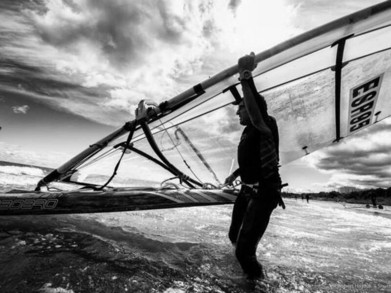 Un windsurfista camina por la playa con su vela, bajo un cielo dramático en blanco y negro.