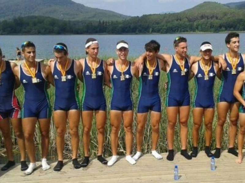 Equipo de remo celebrando una victoria en el lago, con medallas en el cuello y una bandera nacional a su lado.