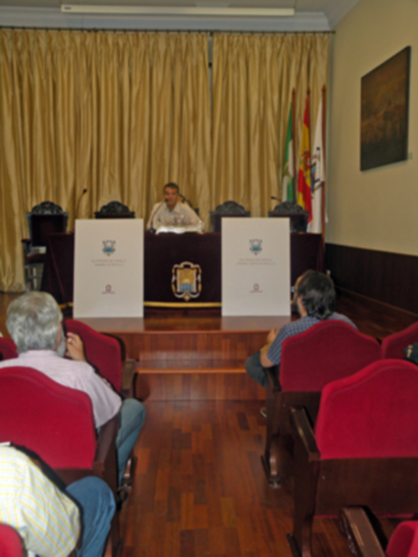 Sala de conferencias con dos personas sentadas en sillas rojas, un hombre hablando en el podium y una bandera en el fondo.