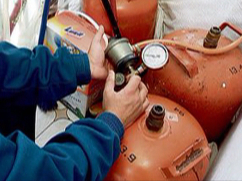 Un individuo ajustando un regulador de gas en un cilindro de gas, con una caja de limpiaplatos en el fondo. El regulador tiene un manómetro para medir la presión del gas.