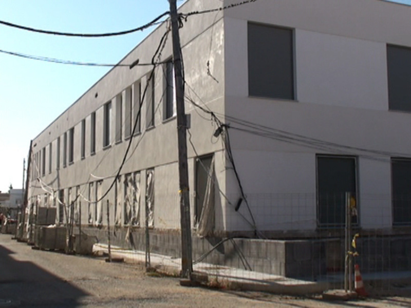 Fachada de un edificio en construcción con cables eléctricos y una cerca de protección en la entrada. El cielo está claro y azul.
