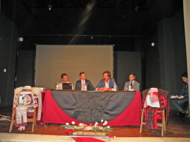 Panel de discusión en un evento formal, con cuatro participantes sentados en una mesa adornada con flores y mantel rojo.