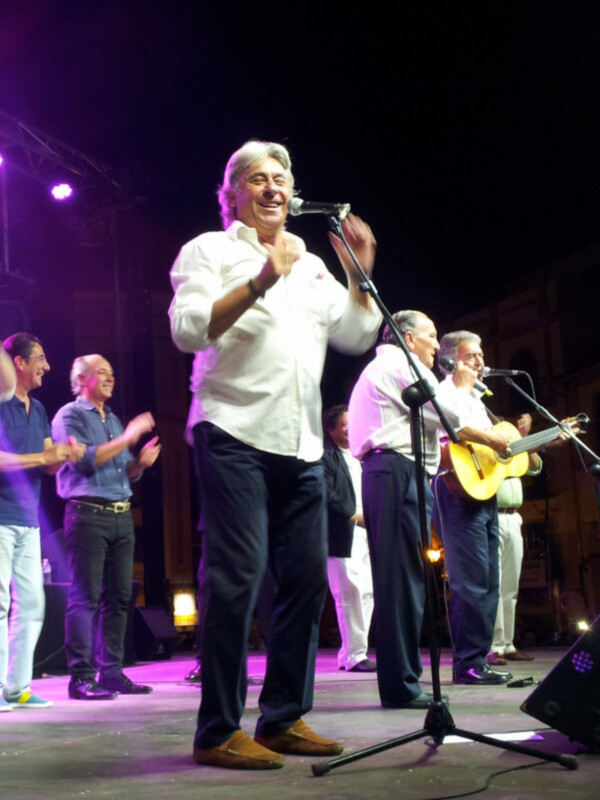 Un músico en un escenario de concierto, cantando con entusiasmo y acompañado por otros músicos.