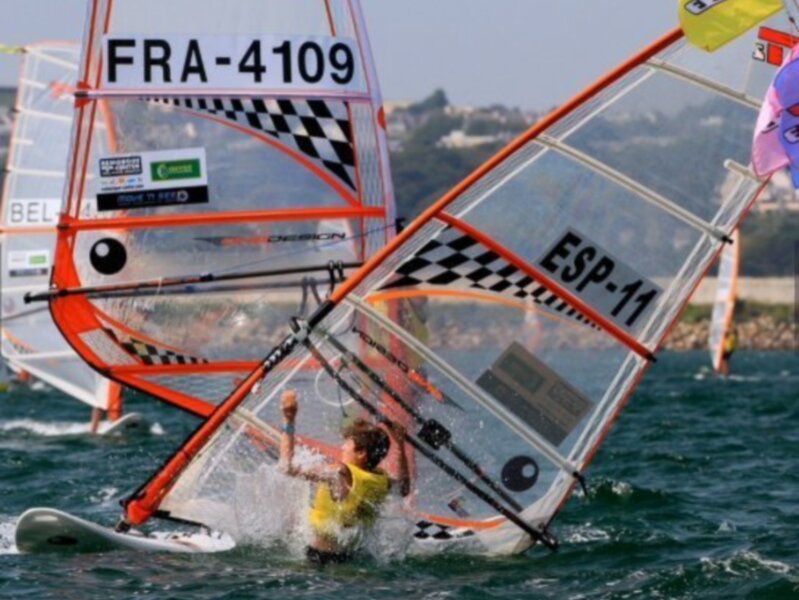 Competidor en una carrera de vela a vela en el agua, con vela española (ESP) y francesa (FRA) en el fondo.