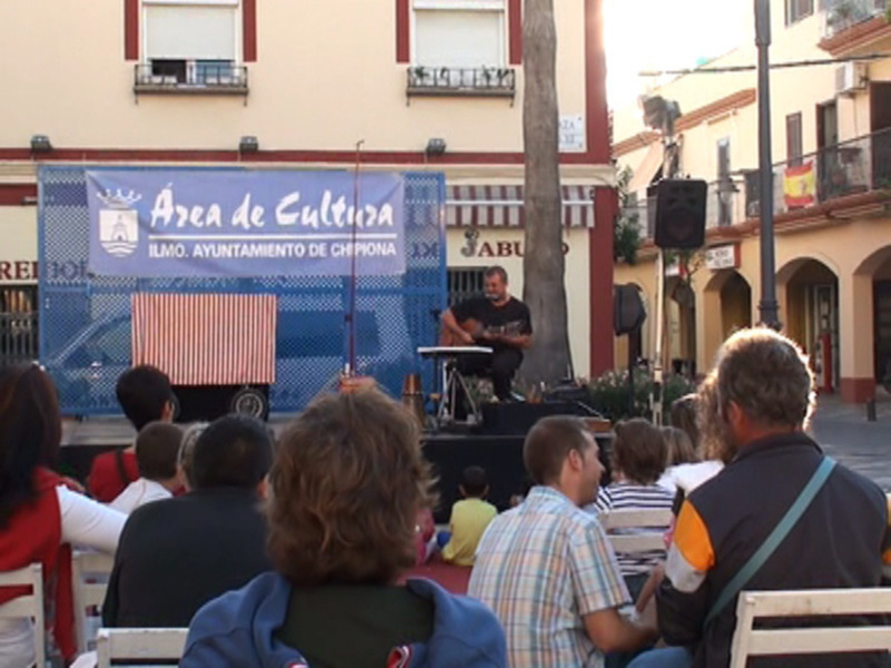 Un grupo de personas disfruta de un concierto en una calle de Chipiona, con un cartel de la 'Área de Cultura' y una bandera americana en el escenario.