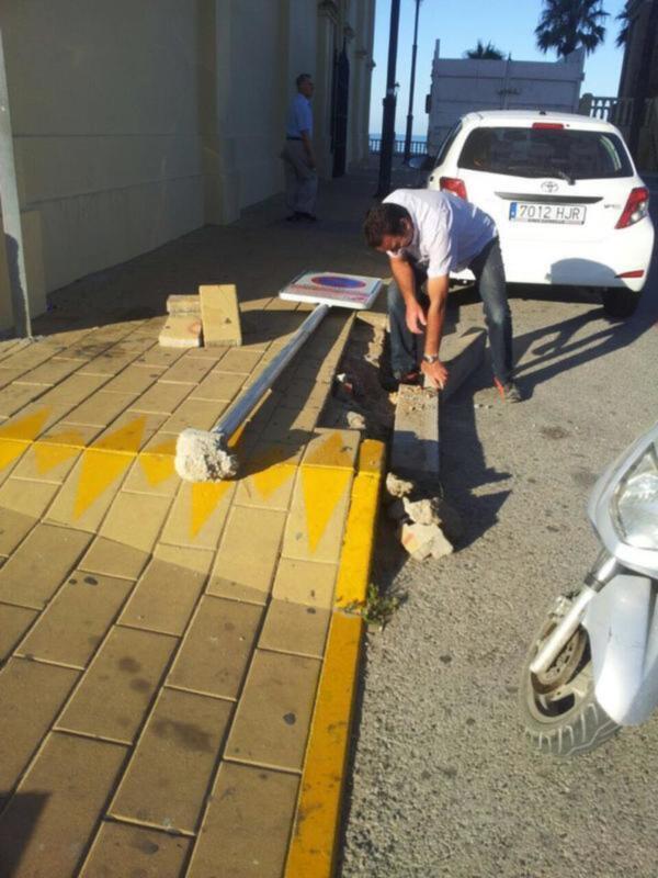 Trabajador reparando una señal de tráfico en una acera junto a un coche estacionado.