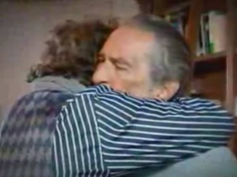 Un hombre con cabello gris y barba está abrazando a una mujer en un sofá. La escena parece ser en un hogar, con estanterías y libros al fondo.