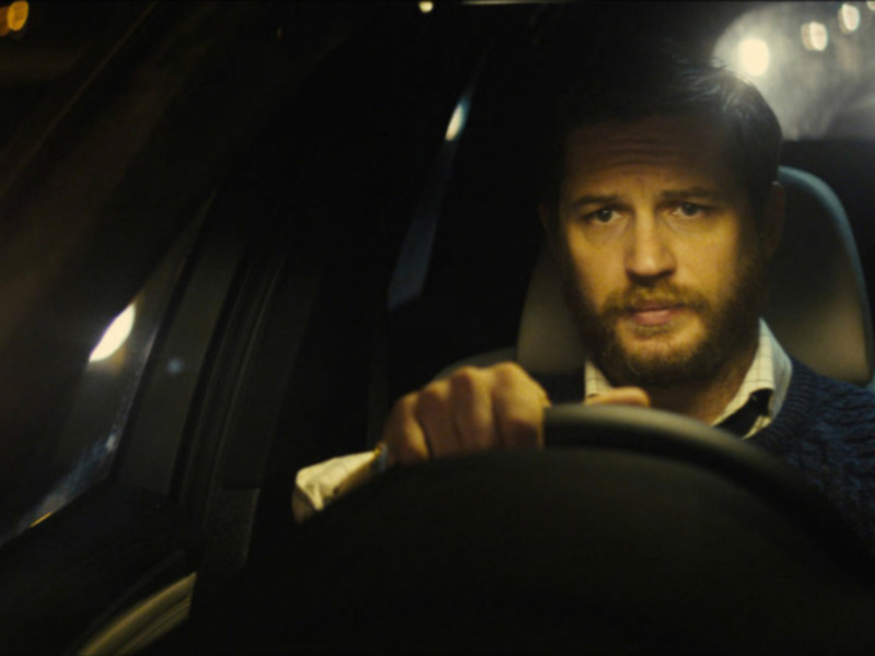 Un hombre con barba y suéter de punto, conduciendo un coche de noche con luces de fondo.