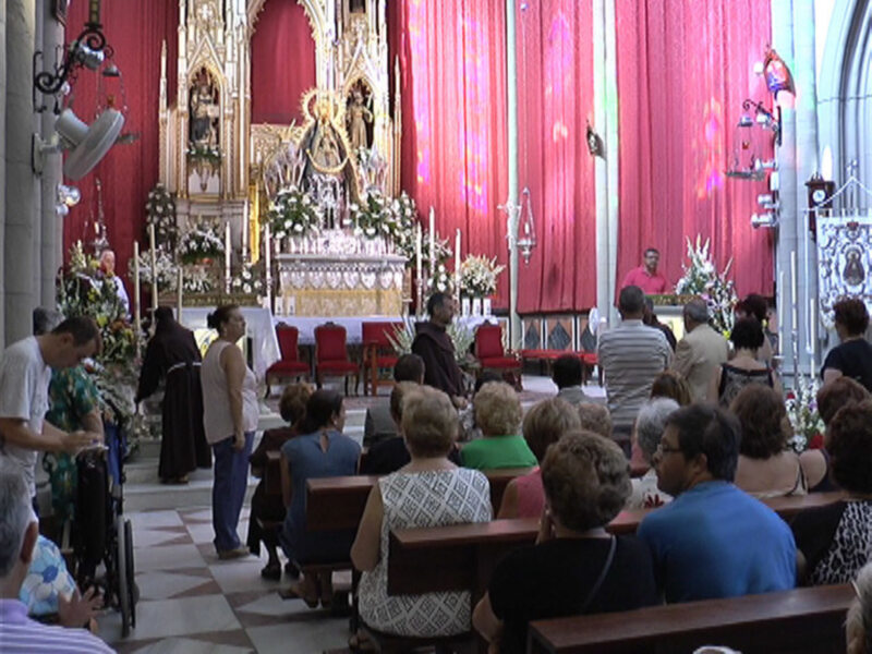 Interior de una iglesia con personas asistiendo a una ceremonia religiosa, con altares decorados y personas sentadas en bancos.