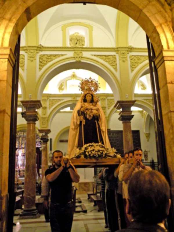 Una imagen de una iglesia con una estatua de la Virgen María en el centro, rodeada por personas que la están protegiendo con sus manos. La arquitectura es elegante con columnas y un techo alto.