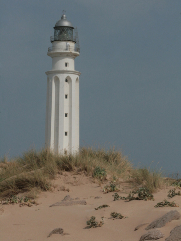 Faro blanco en la costa, rodeado de dunas de arena y vegetación seca.