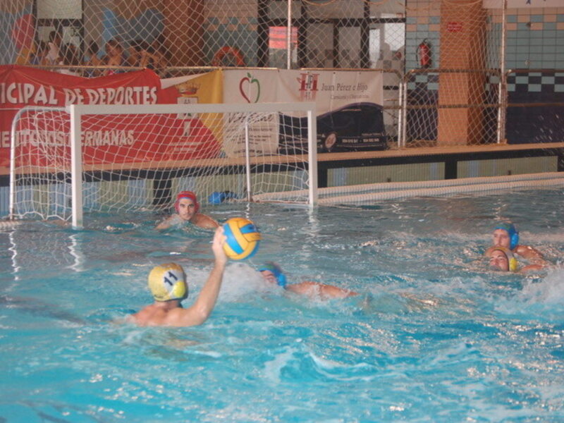 Jugadores de waterpolo en acción en una piscina, con una portería en el fondo y anuncios de clubes deportivos en el muro de fondo. El agua es clara y azul, y los jugadores llevan cascos para protegerse durante el juego.