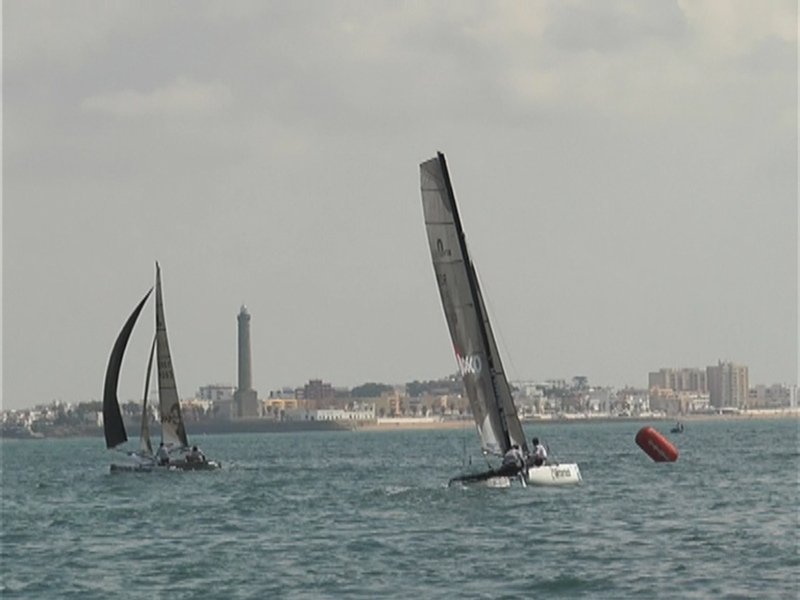 Competición de vela en el mar con barcos navegando cerca de una torre y edificios en el horizonte.