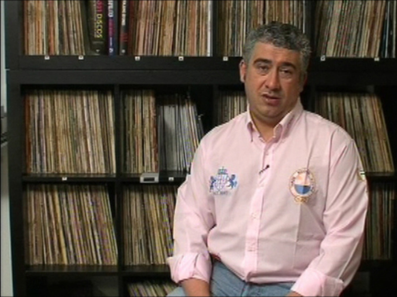 Un hombre sentado frente a una estantería llena de discos vinilo, con una camisa rosa y una insignia en el pecho. La estantería está organizada con discos de diferentes colores y tamaños, creando un fondo visualmente interesante.