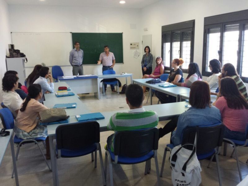 Clase de idioma en un aula con estudiantes sentados en mesas y un profesor frente a una pizarra verde explicando algo en el aula.