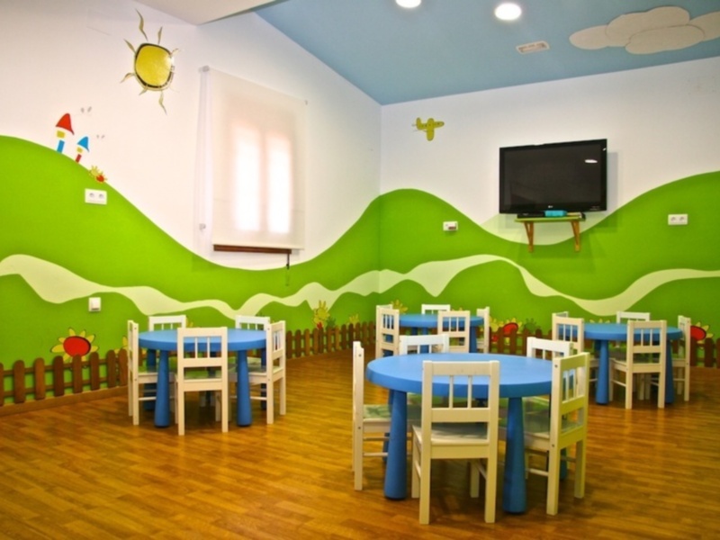Un aula infantil con paredes pintadas con un paisaje de montañas verdes y nubes, mesas azules y sillas blancas. En el techo, hay una televisión y una cortina blanca. La decoración incluye un sol y un pájaro en la pared.