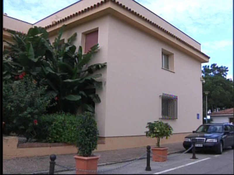 Casa residencial con fachada beige, jardín con plantas y árboles, y coche estacionado en la acera.