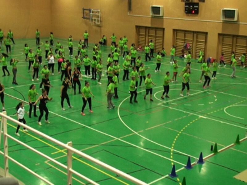 Un grupo de personas en un gimnasio, algunas vestidas de negro y otras de verde, realizando ejercicios de fitness en una pista de baloncesto. Se pueden ver varios conos de entrenamiento en el fondo.