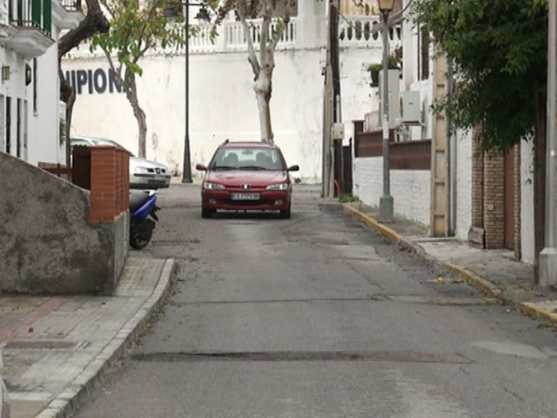 Callejón urbano con coche rojo y motocicleta estacionados, edificios blancos y árboles en el fondo.