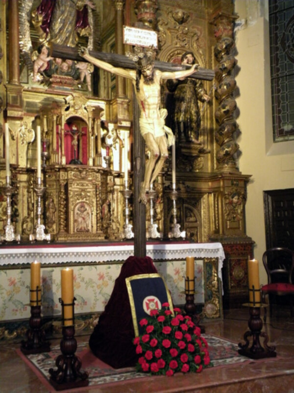 Representación del Cristo crucificado en un altar ornamental y dorado, rodeado de velas y flores rojas.