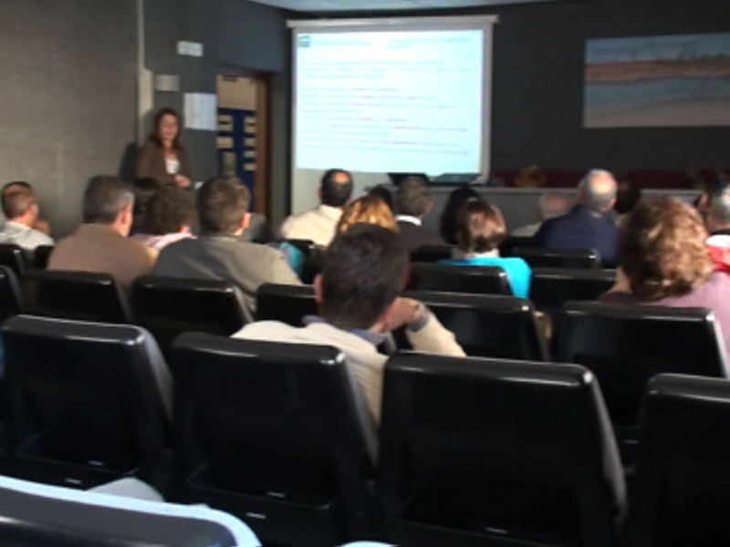 Una sala de conferencias con una pantalla proyectando un presentador y audiencia sentada en sillas.