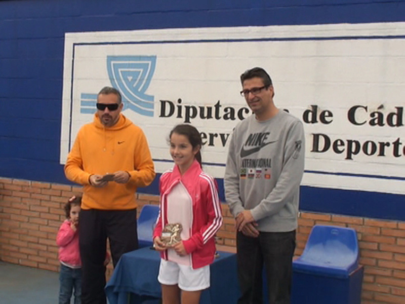 Una niña sonríe mientras sostiene un trofeo junto a dos hombres, uno de ellos sostiene un teléfono móvil. El fondo muestra un cartel con la inscripción 'Diputación de Cádiz'.
