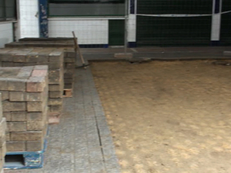 Pavimentación de baldosas en proceso de construcción, con bloques de hormigón a un lado y una estructura de metal en el fondo.